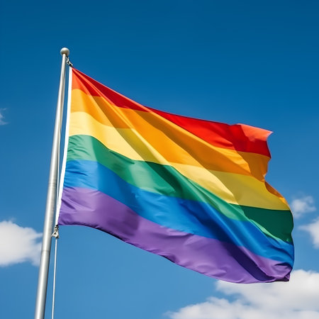Rainbow flag waving in the wind against a blue sky with cloudsの素材