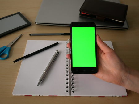 Female hand holding a smartphone with green screen. In the background, office supplies and businessの写真素材