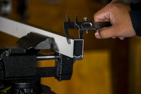 A man using a vernier caliper measuring the width of an aluminum beam thats being held by a bench vise all placed infront of a blurred wooden backgroundの写真素材