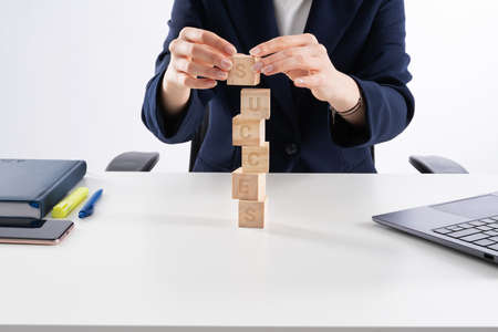 Young caucasian bussines woman stacking wooden blocks in her work office. Office interior, laptop, notebook and flower on white desk.の写真素材