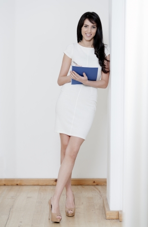 Young woman in white dress leaning against a white wall holding a blue folderの写真素材