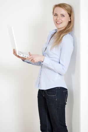 Young woman leaning against a white wall holding a white laptop computerの写真素材
