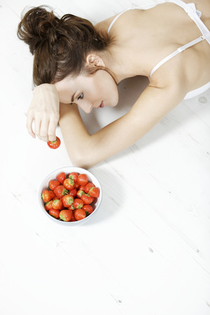 Attractive young woman lying on the floor in her underwear enjoying fresh strawberriesの写真素材