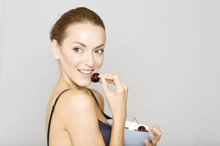 Beautiful young woman in black underwear eating fresh cherries from a bowlの写真素材