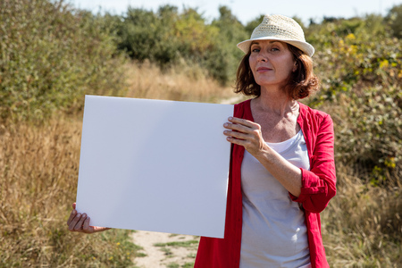 summer outdoors teasing - self-assured 50s woman with seasonal hat holding a white blank ad to write message on board,walking on path in summer grass fieldの写真素材