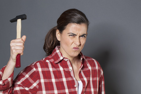 female DIY concept - unhappy young brunette woman holding hammer with violence,losing temper or fed up at having to work manuallyの写真素材