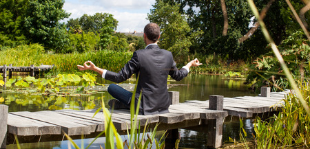 corporate green break - zen middle age male professional practicing yoga on a wooden bridge in the middle of an asian-like green pond for company wellbeing,back view with park foregroundの写真素材