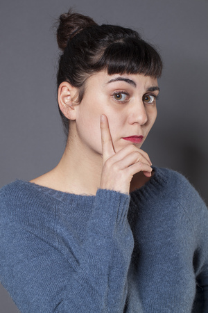 frowning 20s girl with brown hair and blue wool sweater looking surprised with finger on cheek for questionの写真素材