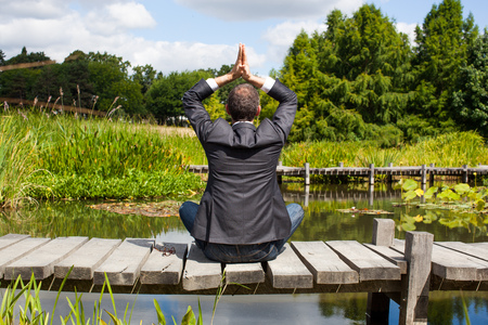corporate green break - zen middle age male professional practicing yoga on a wooden path in the middle of an asian-like green pond for company wellness,back view with park foregroundの写真素材