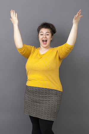 success concept - laughing young overweight girl wearing a yellow sweater expressing her achievement and joy with both hands raisedの写真素材