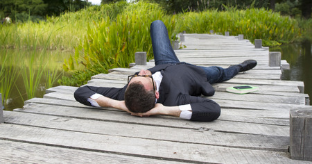 Caucasian modern businessman with hands behind head lying on wood bridge daydreaming and relaxing in city park to think and enjoy lunchtime, back view in daylightの写真素材