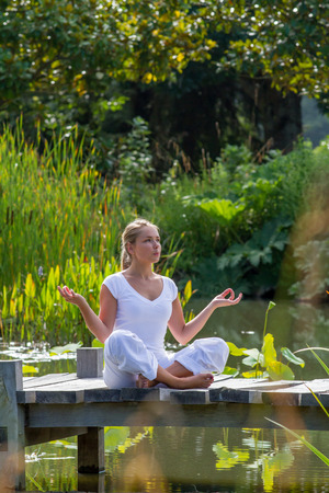 outdoors relaxation - focused young yoga woman in lotus pose,breathing and meditating on a wooden path with green foreground and water lily background, summer exotic daylightの写真素材