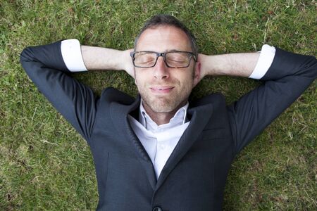 Caucasian modern businessman with grey suit and cool eyeglasses lying on grass sleeping and relaxing with hands behind head in city park enjoying time,overhead view,from above or high angle viewの写真素材