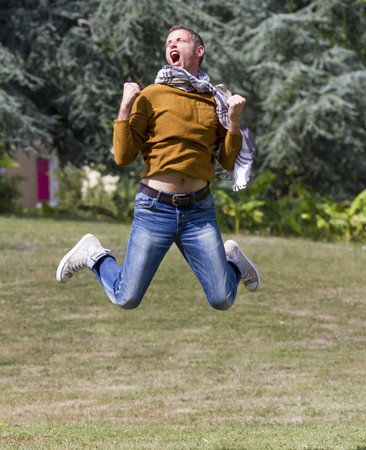 celebrating success outdoors concept - dynamic modern man jumping with arms raised with strength for achievement and happiness in green park,natural summer daylightの写真素材
