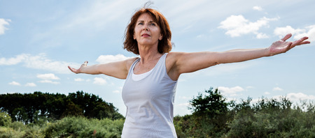 senior zen - radiant mature woman opening her chakra and arms wide,seeking for energy and harmony with nature, green foreground and blue sky in summer daylightの写真素材