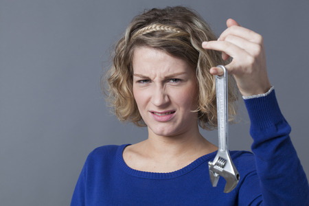 female DIY concept - unhappy young blond woman having disgust at holding spanner as symbol of manual work and mechanics handiworkの写真素材