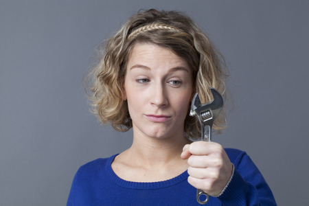 female DIY concept - dubious young blond woman holding wrench,questioning manual work and mechanics handiworkの写真素材