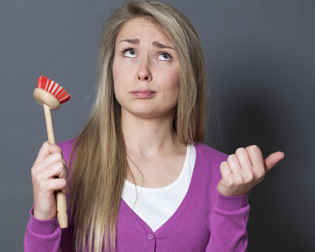 bored young blond woman displaying dish brush for question about washing dishesの写真素材