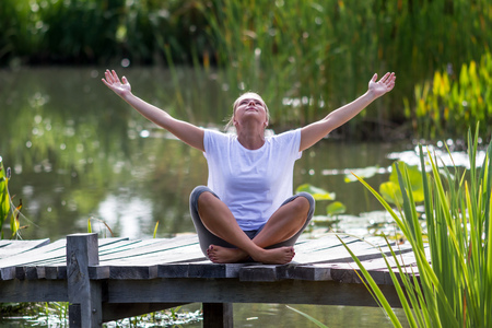 outdoors relaxation - beautiful young yoga woman outstretching her arms,enjoying to relax on a wooden bridge with green foreground and water background, summer daylightの写真素材