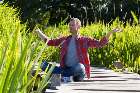 yoga man sitting in lotus position on a wooden bridge with water and reed around breathing and relaxing, seeking for serenity and balance in a park,green environment in summer daylightの写真素材