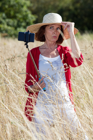 mature selfie concept - focused mature woman learning how to take a selfie with a stick in summer dry meadows wearing a summer hat,natural daylightの写真素材
