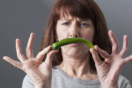 frowning mature woman upset in holding green pepper for spicy aversionの写真素材