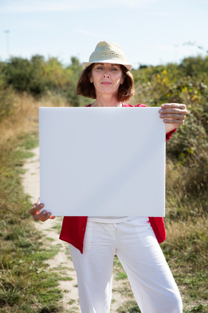 summer outdoors teasing - gorgeous 50s woman with casual hat holding a white blank sign to write message on board,walking on path in summer grass fieldの写真素材