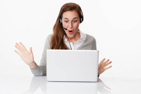 fun at work concept - excited young female manager winning unexpected business with headset on and computer on desk, studio shot,white backgroundの写真素材