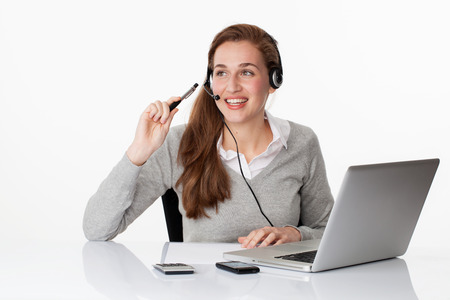 fun at work concept - smiling young female business executive thinking on headset with computer on sparse desk, studio shot,white backgroundの写真素材