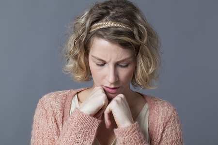 doubt and worry concept - unhappy 20s woman with blond curly hair looking concerned with both fists hiding her chin,grey backgroundの写真素材