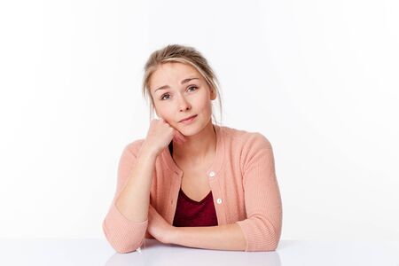 reflection concept - daydreaming beautiful 20s blond woman sitting at a sparse desk for happiness, white background studioの写真素材