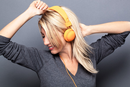 sexy blond girl listening to cool music on headphones, dancing with arms raised for energy, grey background studioの写真素材