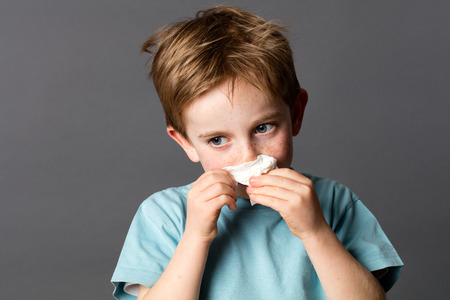 healthcare learning - sick young kid with red hair and freckles using a tissue after a cold, rhinitis or spring allergies, grey background studioの写真素材
