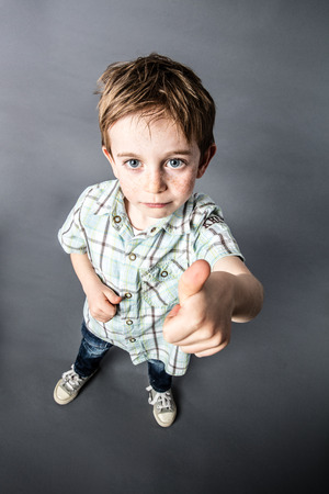 cheeky OK - beautiful little red hair boy with freckles expressing his satisfaction, approval and mischievous success with thumbs up in the foreground with wide angle and top view, contrast effects, grey background studioの写真素材