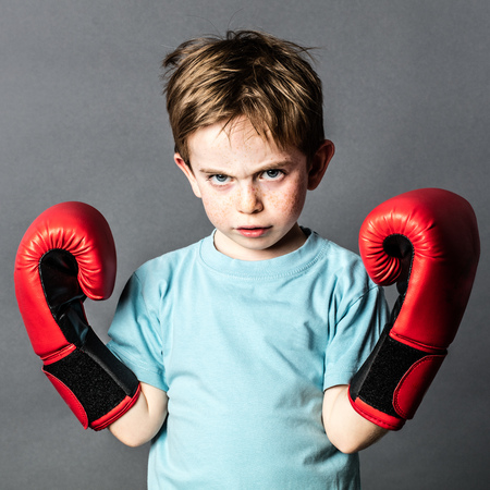 unhappy beautiful young male preschooler with freckles and red hair showing his boxing gloves up, sulking or looking aggressive for sport, grey background studioの写真素材