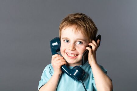 cheeky young red hair boy talking on an old fashioned telephone and a smartphone on both ears for two voices communication, grey background studioの写真素材