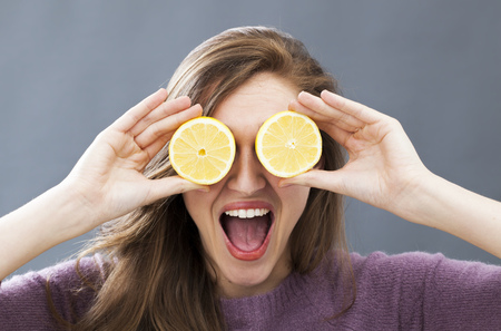 laughing beautiful young woman with two zesty lemon slices on her eyes for fresh skincare and vegetarian wellbeing, indoorsの写真素材