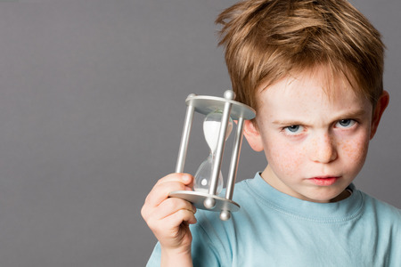 unhappy little boy with red hair showing his impatience with an egg timer in his hands for time...