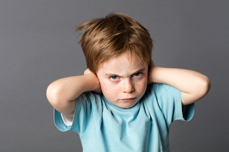 stubborn little kid with red hair, freckles and an attitude ignoring parents scolding, blocking his ears with hands against education problems, grey backgroundの写真素材