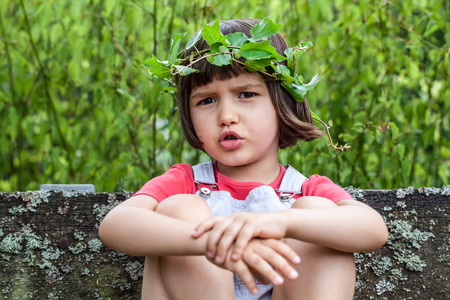 portrait of an unhappy young child with a crown made of green ivy leaves sitting on a mossy bench, relaxing in garden in summertimeの写真素材