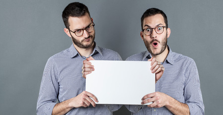 concept of confusion in communication with young businessman with eyeglasses showing a copy space banner expressing positive and negative emotions, indoorsの写真素材