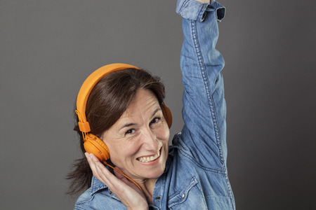 fun middle aged woman listening to music on orange headphones, raising her arms for happiness and...