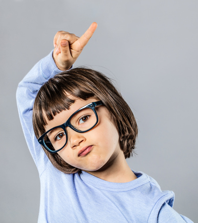 unsure beautiful little child with serious eyeglasses raising her finger to ask or answer a question at school for learning and education idea, grey backgroundの写真素材