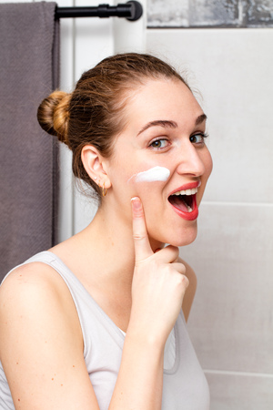 laughing beautiful girl enjoying applying moisturizing face cream on her cheek for fun morning routine in her bathroomの写真素材