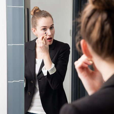 focused beautiful young smart woman with a jacket applying her eye concealer in front of her bathroom mirror for makeup morning routineの写真素材