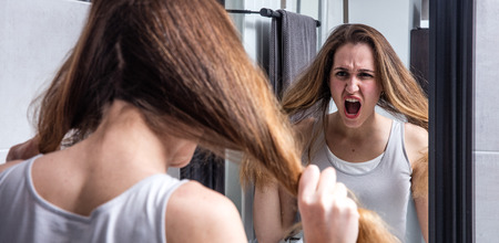 young woman in crisis shouting in front of her mirror, pulling out her split tousled long hair for stressed out haircare and beauty, real people in home bathroomの写真素材