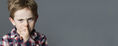 unhappy young boy with red hair sulking with a finger on lips for secret or severe teacher-like gesture, grey copy spaceの写真素材