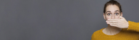 negative feelings concept - portrait of amazed beautiful 20s girl covering her mouth for gag metaphor,studio shot on long gray copyspaceの写真素材