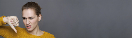 negative feelings concept - portrait of grumpy 20s girl disapproving with thumbs down,studio shot on gray copy space panoramaの写真素材