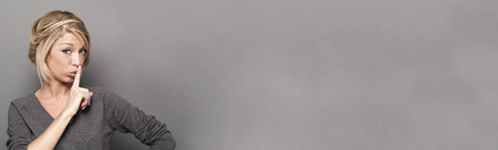 fun secret concept - sexy young blond woman requiring discretion with a finger on her lips,studio shot, grey copy space long bannerの写真素材